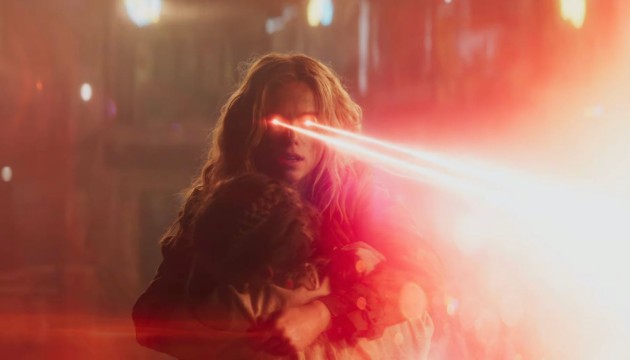 Supergirl segura uma criança nos braços enquanto dispara seus raios de calor, iluminando a cena com um intenso feixe vermelho.
